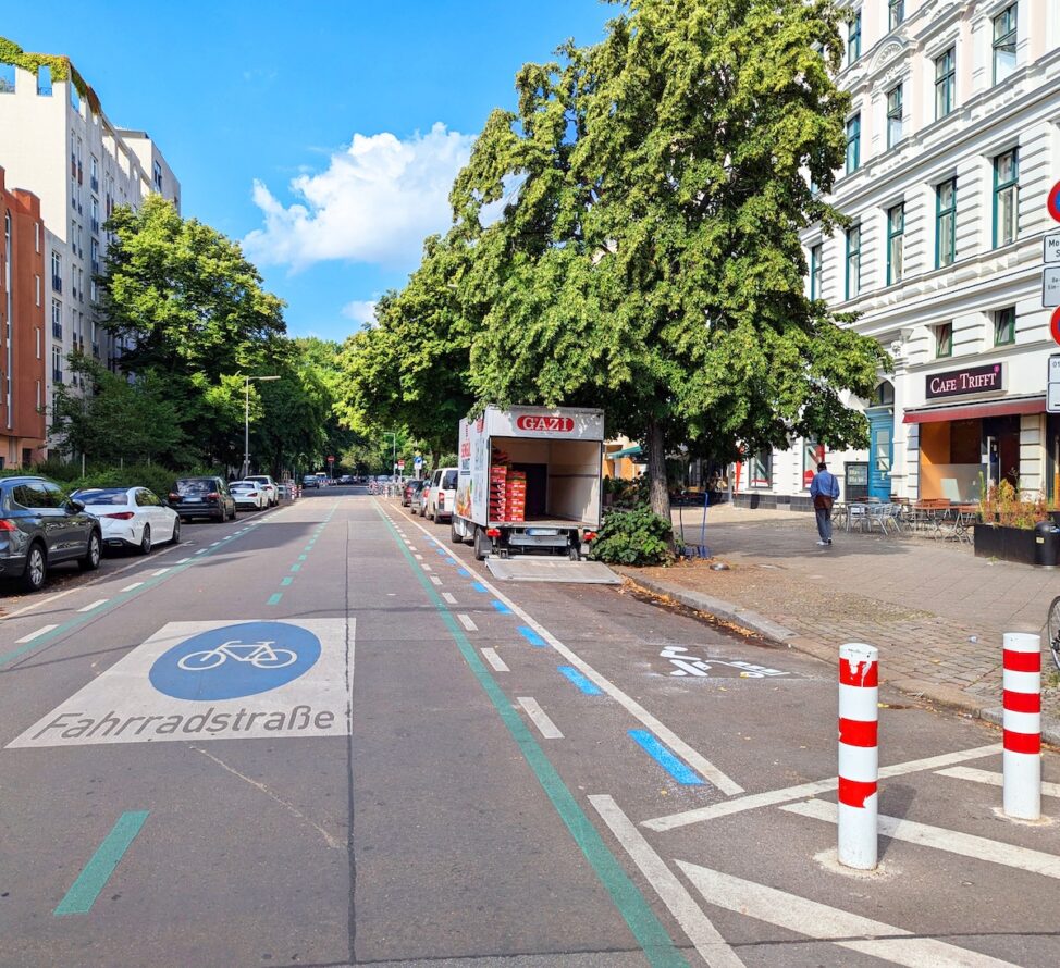 Straße mit Fahrzeugen und Bäumen, Verkehrszeichen. Es ist eine Fahrradstraße und ein Ladebereich ist zu sehen.
